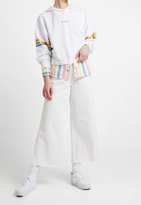 Felpa bianca corta con strisce arcobaleno sulle maniche, cinte in vita. Abbinata a pantaloni in denim bianchi a gamba larga e sneaker bianche.