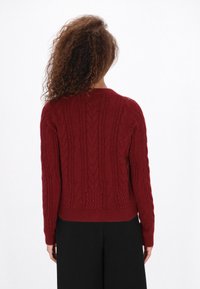 Göndör hajú nő piros kötött pulóverben és fekete nadrágban, háttal állva egy sima fehér háttér előtt.