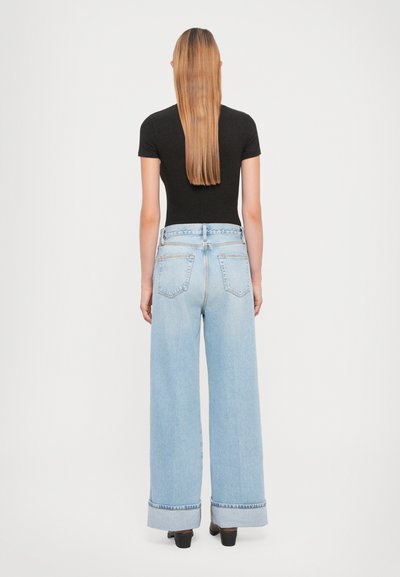 Woman with long straight hair wearing a black short-sleeve top, high-waisted wide-leg light blue jeans, and black heeled shoes, standing facing away.