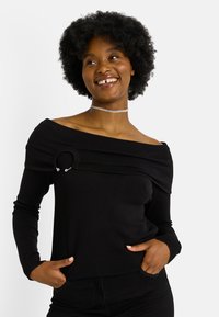 Mujer sonriente con cabello afro natural corto lleva un suéter negro con hombros descubiertos y detalle de anillo, además de un collar choker de plata, con las manos en los bolsillos.