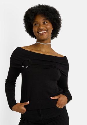 Femme souriante avec des cheveux afro naturels courts portant un pull noir à épaules dénudées avec un détail en anneau et un collier en argent, les mains dans les poches.