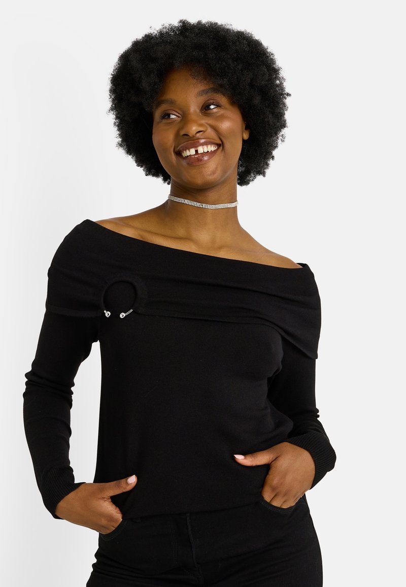 Mujer sonriente con cabello afro natural corto lleva un suéter negro con hombros descubiertos y detalle de anillo, además de un collar choker de plata, con las manos en los bolsillos.