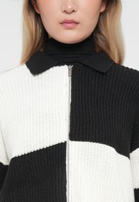 Sweater em malha waffle com um design em blocos de cor preto e branco, apresentando um colarinho e um fecho éclair frontal. Tecido texturizado com seções contrastantes.