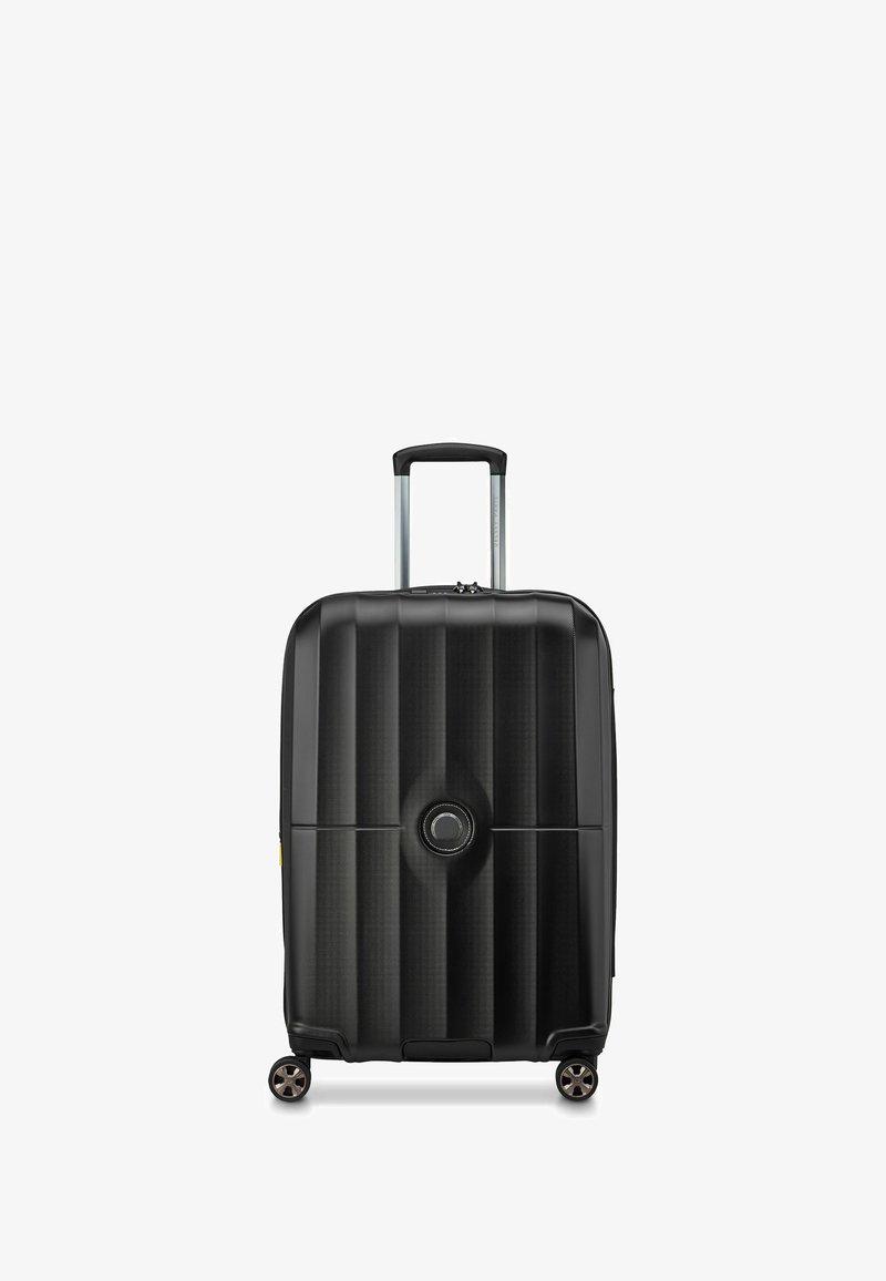 Valise rigide noire à rainures verticales, poignée télescopique argentée, quatre roues et un logo circulaire sur le devant.