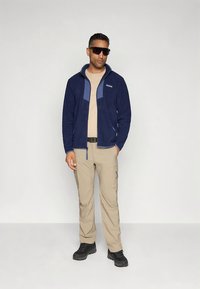 Marineblauwe fleecejas met ritssluiting aan de voorkant en contrastpanelen, gecombineerd met beige cargobroek en zwarte laarzen. Zonnebril maakt de outfit compleet.