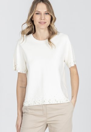 Blondynka w białej bluzce z krótkim rękawem zdobionej koralikami na rękawach i dole, w beżowych spodniach, delikatnie się uśmiecha.