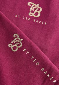 Maroon kangas ambayo ina hisa za dhahabu zikiwa na nembo "TB" na "by Ted Baker." Uso laini wenye mwangaza wa kidogo, ukionyesha herufi na mapambo tofauti.