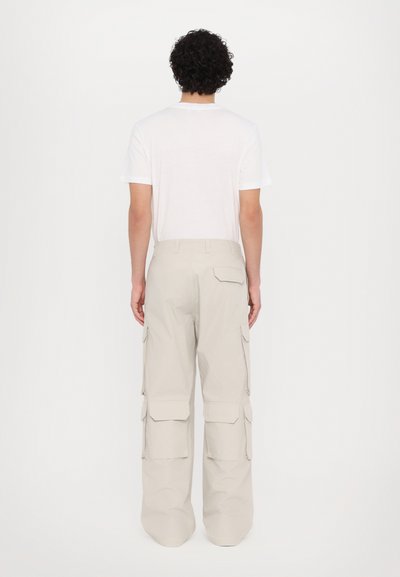 Pantaloni cargo bej, cu mai multe buzunare laterale, croi lejer și design conic, asortați cu un tricou alb simplu, prezentați din spate.