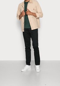 Beige Jacke mit Kragen und zwei Fronttaschen, kombiniert mit einem grünen T-Shirt und schwarzen Hosen, ergänzt durch weiße Sneakers.