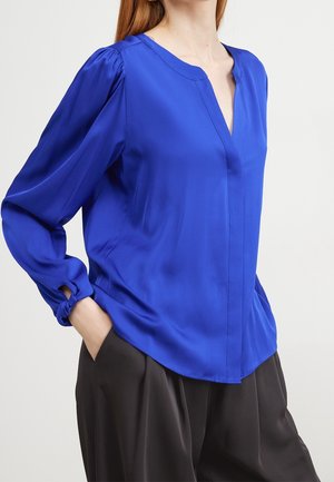 Frau trägt eine leuchtend blaue Bluse mit langen Ärmeln und V-Ausschnitt sowie lockere schwarze Hosen, Hände teilweise in den Taschen, vor einfachem Hintergrund.