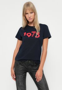 Navy zila kokvilnas t-kreklu ar drosmīgu sarkanu "1973" grafiku. Īsas piedurknes un apaļa kakla izgriezums. Kombinēts ar melniem teksturētiem džinsiem.