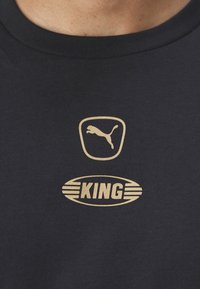 Juodas medvilninis marškinėlis su auksiniu Puma logotipu virš žodžio "KING" ovalo formoje, po kuriuo yra horizontalios juostos.
