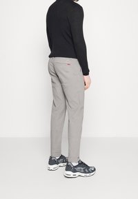 Chino grigi con taglio slim, abbinati a un maglione nero a coste. Accentuati da un piccolo etichetta rossa sul taschino posteriore. Scarpe da ginnastica.