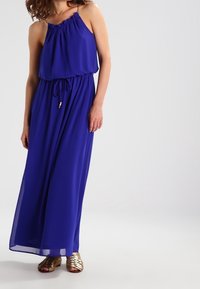 Kungsblå maxi-klänning med en rynkad halsringning, justerbar dragsko i midjan och flödande tyg. Bärs med guldiga sandaletter med remmar.