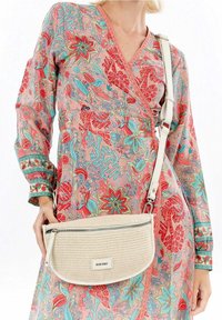 Borsa a tracolla bianca con superficie testurizzata e chiusura a zip, tenuta davanti a un capo d'abbigliamento a fantasia floreale multicolore.