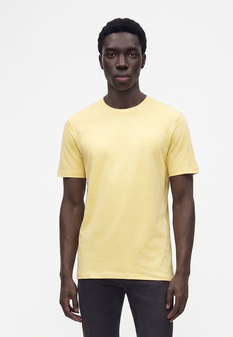 Homme à la peau foncée portant un t-shirt jaune clair uni à manches courtes et un pantalon sombre, debout devant un fond blanc.