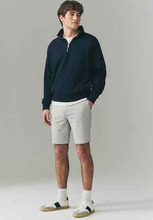 Sudadera de media cremallera en color navy, pantalones cortos grises claros, zapatillas blancas con detalles en negro y calcetines blancos. El modelo está de pie en un fondo neutro.