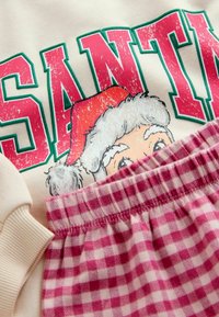 Kerman huppari, jossa on vihreä ja vaaleanpunainen "SANTA" -teksti ja sarjakuvamainen joulupukkikasvo, yhdistettynä vaaleanpunaisiin ja valkoisiin ruudullisiin pyjamahousuihin, joissa on joustava vyötärö.