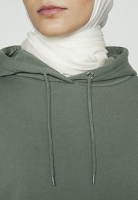 Primer plano de una persona que lleva un hijab blanco y una sudadera con capucha verde, con cordones visibles, contra un fondo liso.