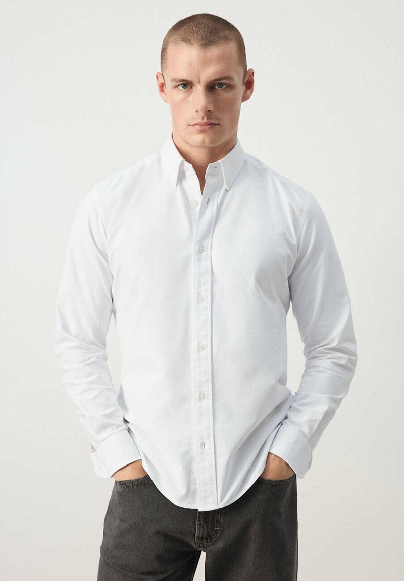 Homme aux cheveux courts portant une chemise blanche boutonnée et un pantalon sombre, debout les mains dans les poches devant un fond clair uni.