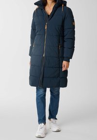 Manteau matelassé long bleu marine avec capuche, présentant une fermeture éclair à l'avant et des accents en matériel beige. Le tissu est matelassé avec une texture rembourrée.