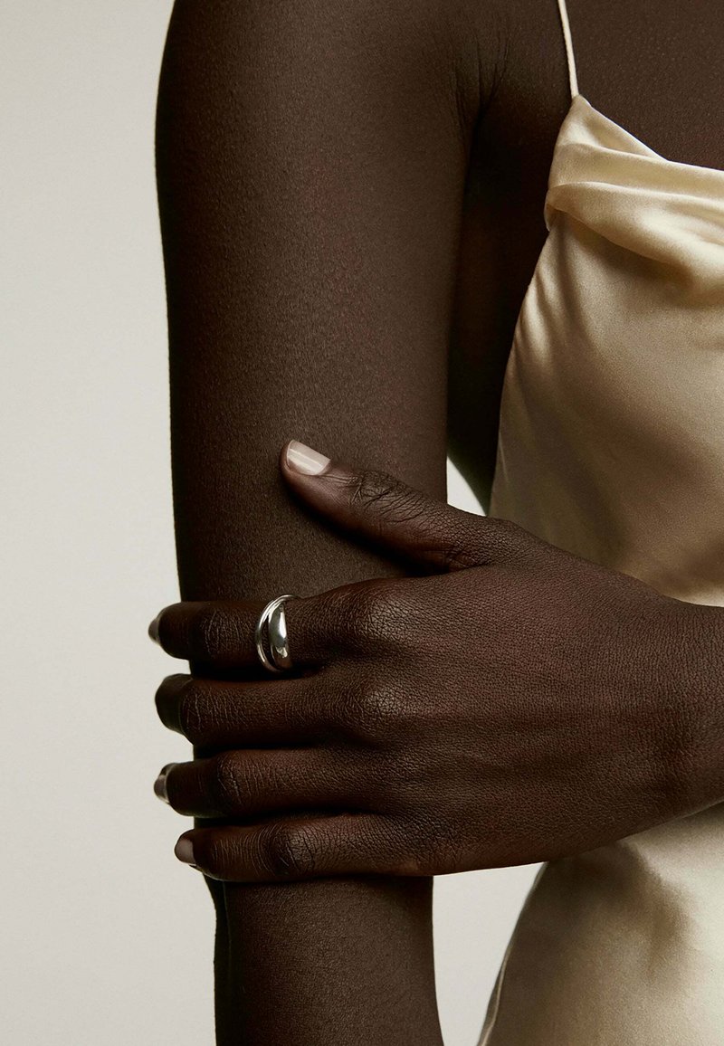 Anillo de plata en una mano con piel oscura texturada, reposando sobre un brazo. El fondo presenta un tono claro y liso, que complementa la tela satinada.