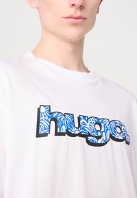 White cotton t-shirt featuring a stylized "hugo." logo in blue with a wave-like pattern, outlined in black. Casual fit with a crew neckline.