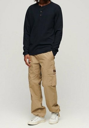 Mand iført navyblå langærmet henley-shirt, beige cargobukser med sidelommer og hvide sneakers, stående mod en ensfarvet baggrund.