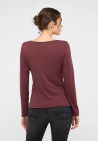 Langärmeliges, kastanienbraunes Hemd mit weitem Ausschnitt. Aus weichem Stoff gefertigt, mit kleinem Logo auf der Rückseite, kombiniert mit dunkler Jeans.