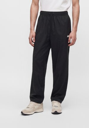 Mand iført sorte sportbukser med elastisk talje og beige sneakers, stående mod en ensfarvet lys baggrund.