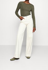 Groen, langmouw fitted top met een subtiele glans, gecombineerd met hoge taille, wijdvallende witte broek en groene schoenen.