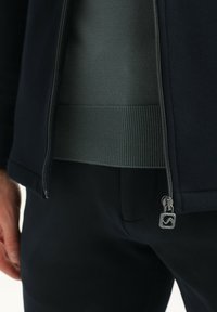 Chaqueta de punto oscuro con cremallera sobre una sudadera verde claro con ribetes. Cuenta con un tirador de cremallera de metal con detalle texturizado. Acabado de tela suave.