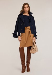 Frau in einer marineblauen Jacke über einem marineblauen Oberteil, einem beigen, taillierten Rock mit Taschen, braunen knietiefen Stiefeln und einer beigen Handtasche.