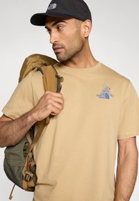 Μπεζ μπλουζάκι με μπλε λογότυπο στο στήθος, φορεμένο με καφέ και πράσινο σακίδιο πλάτης με ρυθμιζόμενους ιμάντες και αγκράφες. Μαύρο καπέλο ολοκληρώνει την εμφάνιση.