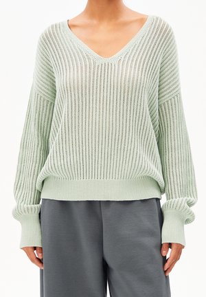 Kobieta ubrana w jasnozielony dziergany sweter z dekoltem w serek, z prążkowanymi mankietami i szare luźne spodnie.