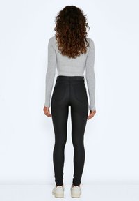 Femme aux cheveux bouclés portant un haut gris à manches longues, un pantalon skinny noir taille haute et des baskets blanches, debout de dos.