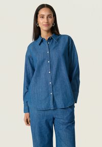 Cămașă din denim de culoare bleu deschis, cu mâneci lungi, închidere cu nasturi și guler ascuțit. Purtați cu pantaloni din denim asortați. Textură netedă.