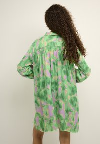 Oversized Hemdkleid mit grünem und lila Camouflage-Muster, satinierter Textur, langen Ärmeln, gesenkten Schultern und gerafftem Rückendetail.