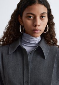 Gray, textured button-up shirt with a collar, layered over a light gray turtleneck. Model wears large silver hoop earrings.