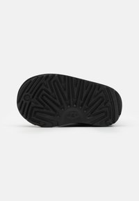 Semelle en caoutchouc noir d'une chaussure présentant un motif zigzag texturé et un logo en relief. Bords lisses et arrondis sans fixations visibles.