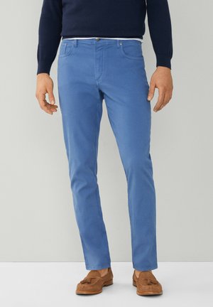 Jeans en coton bleu vif avec une coupe slim, présentant une fermeture éclair, cinq poches et des détails de couture subtils. Portés avec des mocassins marron.