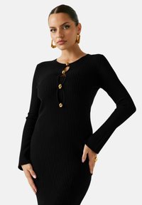Sort ribstrikket kjole med tætsiddende silhuet, der har en rund halsudskæring, lange ærmer og store guldknapper langs forsiden.