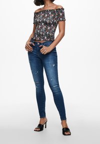 Off-shoulder kukka-toppi, jossa on rypytetty yksityiskohta, yhdistettyinä kuluneisiin sinisiin skinny-farkkuihin ja mustiin avonaisiin korkokenkiin.