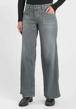 Femme portant un jean gris à jambes larges avec de grandes poches avant et des mocassins noirs, main dans la poche, sur un fond uni.