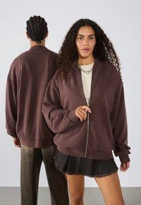 Smeđa oversized bomber jakna sa zipom i rebrastim manžetama, uparena s bež majicom i crnom plisiranom traper suknjom, modeliraju je dvije osobe.