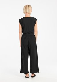 Zwarte jumpsuit met korte fladdermouwen, losse pasvorm, textiel stof, wijde pijpen en een ceintuur om de taille. Model wordt van achteren getoond.