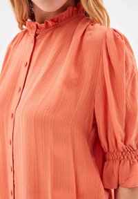 Blouse couleur pêche avec des rayures verticales texturées, des manches bouffantes plissées et un col à volants avec des fermetures à boutons sur le devant.