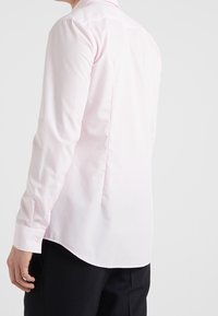 Camisa de manga larga rosa claro con cuello puntiagudo, canesú en la parte posterior de los hombros y dobladillo curvo, confeccionada en tela de algodón suave con puños abotonados.
