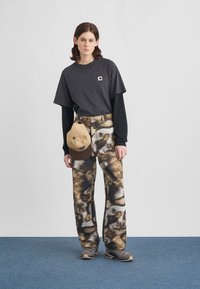Black oversized t-shirt with a logo patch, brown patterned trousers, and a beige cap. Gray sneakers complete the outfit.