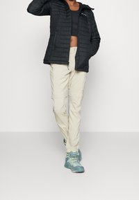 Giacca a vento nera imbottita con tasche con zip, pantaloni cargo beige chiaro con tasche cargo e scarpe da trekking blu con lacci e suole texture.